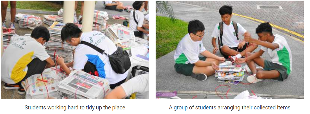 Students in white uniforms bundling stacks of newspapers with string outdoors on the ground.